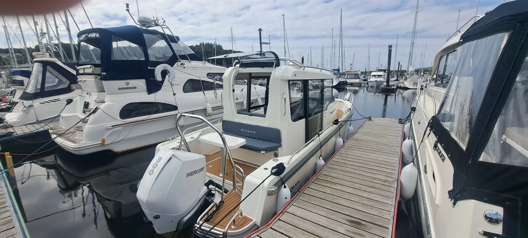 A white motorboat with a Mercury 300 engine is docked at a marina alongside other boats, with a wooden walkway visible.